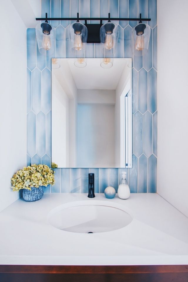 Blue tile behind bathroom mirror with white countertop and dark light fixture 